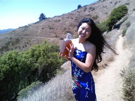 Rana Chang, owner and operator of House Kombucha, holds a growler of her beverage while standing on a hillside path in a blue dress.