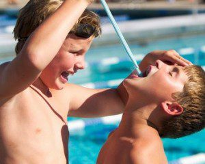 Pikie Sticks for Kids A smiling child pours the contents of a blue Pixy Stick into the open mouth of another child while standing next to a pool.