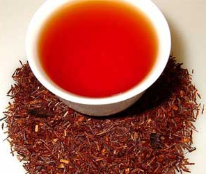 A cup of redbush rooibos tea sits atop a pile of roasted honeybush leaves and stems.