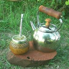 A gourd and bombilla, metal straw, used for drinking yerba mate sits atop a stone next to a kettle.